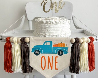 May include: A blue truck with a white cab and bed, carrying three orange pumpkins, is on a white banner with the word "ONE" in orange letters. The banner is strung with brown, white, and orange tassels. A white cake with white frosting is on a white high chair behind the banner.