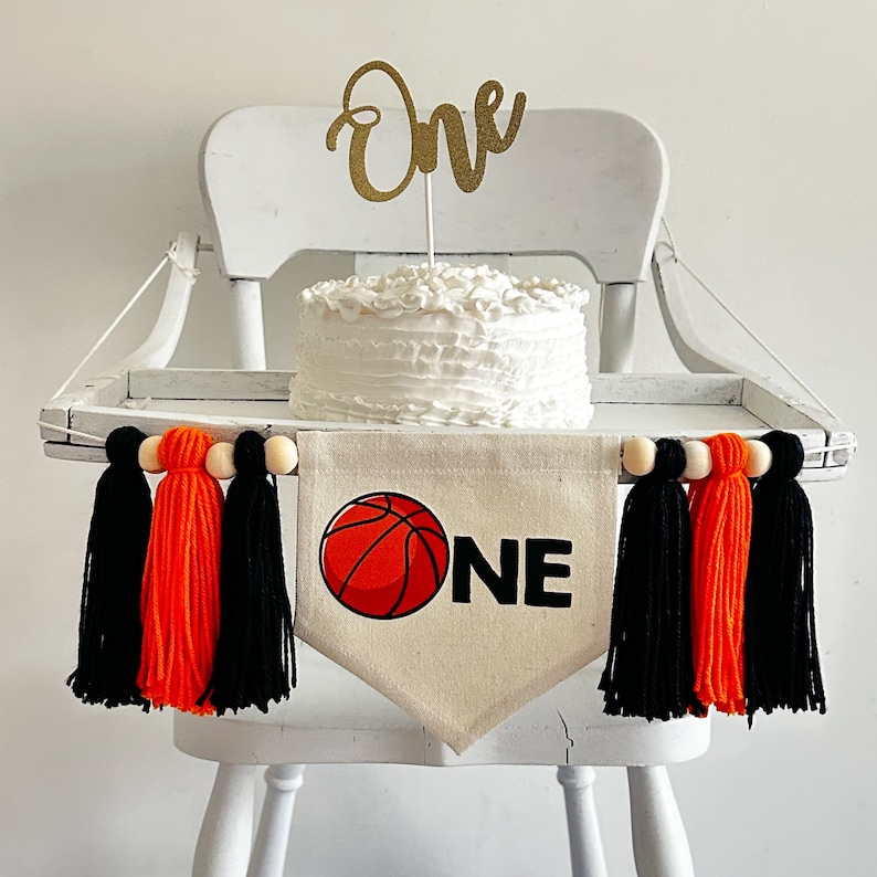 May include: A white high chair with a white cake decorated with white frosting and a gold glitter "One" topper. A banner with a basketball graphic and the word "ONE" hangs from the chair. The banner is decorated with black and orange tassels.