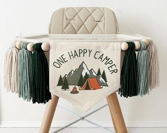 Könnte beinhalten: Ein cremefarbenes Stoffbanner mit dem Text ONE HAPPY CAMPER über einer Grafik aus Bergen, Tannen und einem orangefarbenen Zelt mit Lagerfeuer. Das Banner ist mit Holzperlen und Garnquasten in Grün- und Beigetönen verziert.