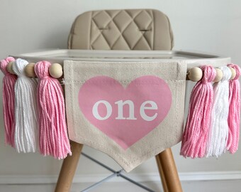 May include: A pink and white high chair banner with the word "one" in a pink heart. The banner is made of fabric with wooden beads and tassels.