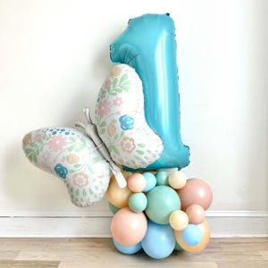 Può includere: Un palloncino blu a forma di numero uno con un palloncino a farfalla floreale attaccato alla parte inferiore. I palloncini sono circondati da un gruppo di palloncini color pastello.