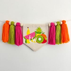 May include: A colorful fruit-themed banner with the word "two" in white letters. The banner is made of fabric and has a triangular shape. It is decorated with a watermelon, pineapple, kiwi, banana, and apple. The banner is strung with wooden beads and colorful tassels.