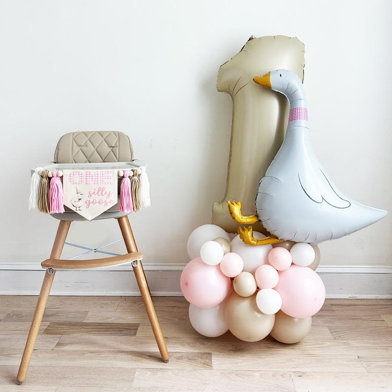 May include: A high chair with a banner that reads "ONE silly goose" and a balloon arrangement. The balloon arrangement includes a large number one balloon, a goose balloon, and a cluster of pink, white, and tan balloons.