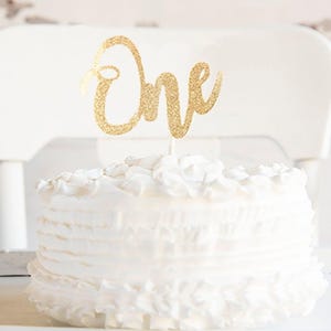 A white cake with a gold "One" topper sits on a white table, with a white chair visible in the background.