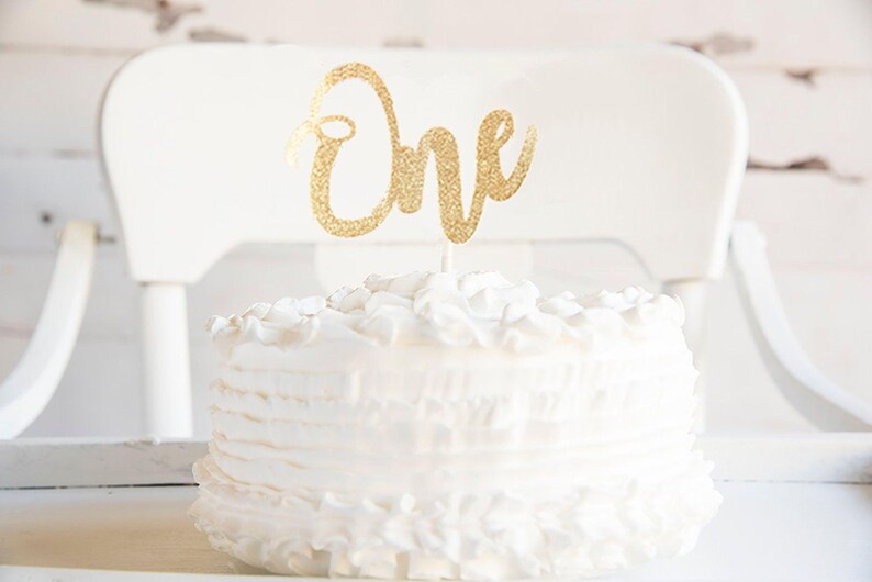 A white cake with a gold "One" topper sits on a white table, with a white chair visible in the background.