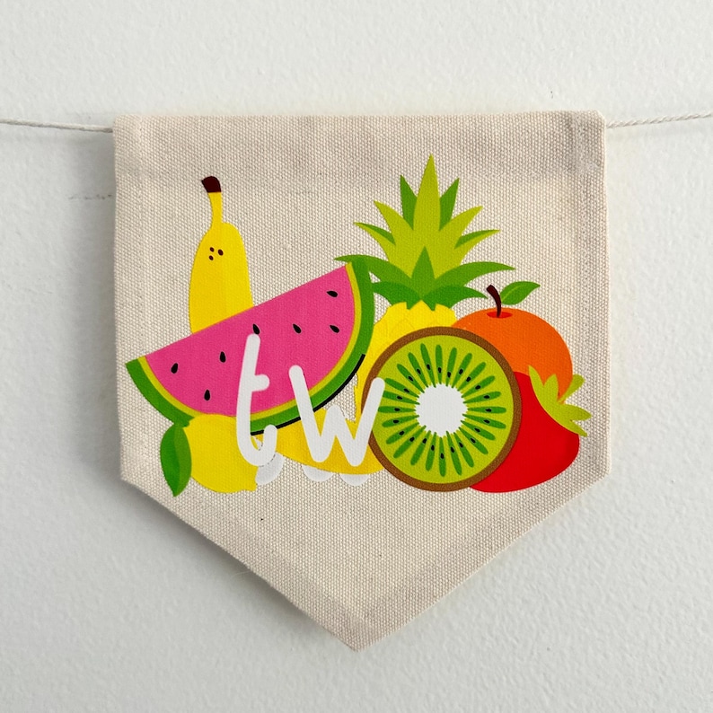 May include: A white canvas banner with a pocket shape at the bottom. The banner features a colorful fruit design with a watermelon, kiwi, banana, pineapple, and a red strawberry. The word "TWO" is written in white letters on the banner.