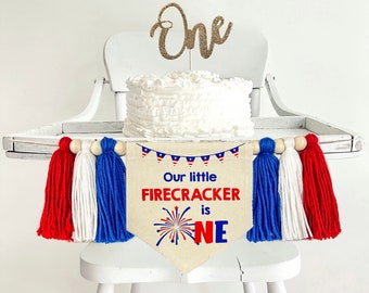 May include: A white high chair with a white cake on top. The cake has a gold glitter "One" topper. A banner with red, white, and blue tassels hangs from the back of the chair. The banner says "Our little firecracker is one" with a red, white, and blue firework graphic.