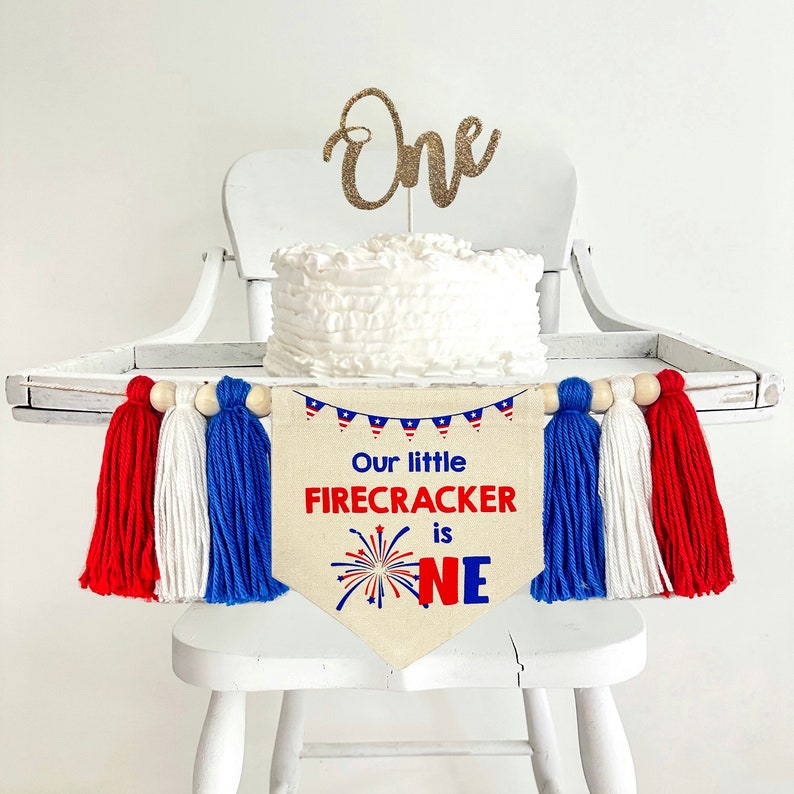 May include: A white high chair with a white cake on top. The cake has a gold glitter "One" topper. A banner with red, white, and blue tassels hangs from the back of the chair. The banner says "Our little firecracker is one" with a red, white, and blue firework graphic.