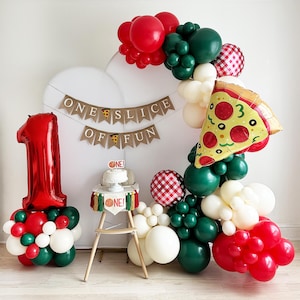 May include: A festive first birthday party setup with a pizza theme. Features a large red number one balloon, a balloon arch with red, green, and white balloons, and a pizza slice balloon. A banner reads "ONE SLICE OF FUN."