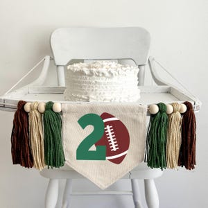Puede incluir: Una silla alta blanca con un pastel blanco en la parte superior y una pancarta verde y marrón con el número "2" y una pelota de fútbol. La pancarta tiene borlas marrones, beige y verdes.