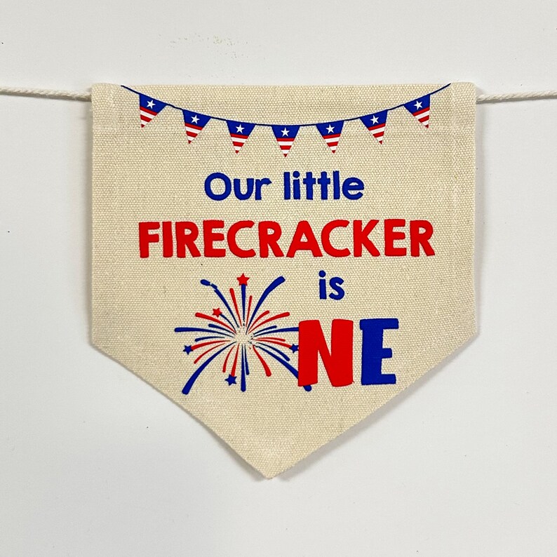 May include: A beige fabric banner with a red, white, and blue American flag garland at the top. The banner says "Our little FIRECRACKER is ONE" with a red and blue firework graphic and the number one in red and blue.