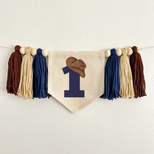 May include: A beige fabric banner with a blue number one and a brown cowboy hat on top. The banner is decorated with brown, beige, and blue tassels.