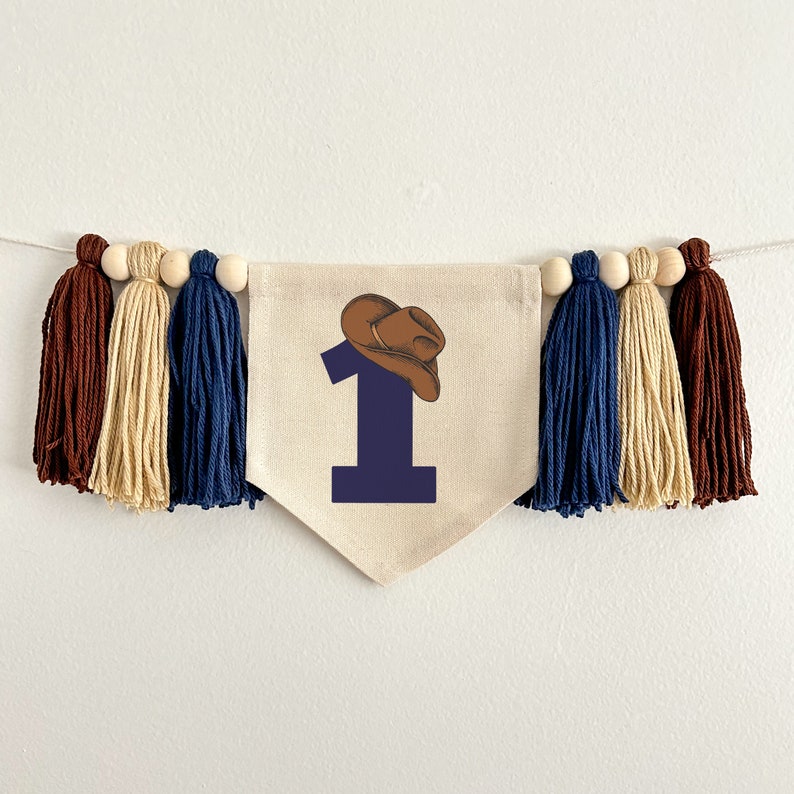 May include: A beige fabric banner with a blue number one and a brown cowboy hat on top. The banner is decorated with brown, beige, and blue tassels.
