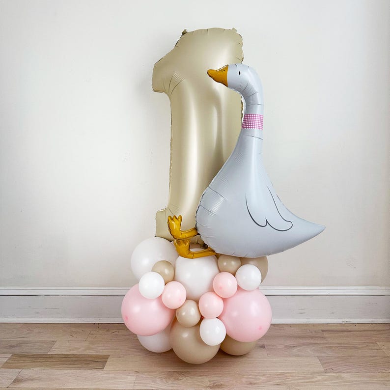 May include: A gold number one balloon and a gray goose balloon are atop a cluster of pink, white, and tan balloons. The goose has a pink and white checkered collar and a yellow beak. The balloons are arranged for a first birthday celebration.