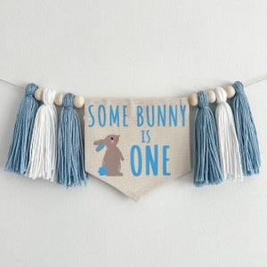 May include: A blue and white tassel garland with a banner that reads "Some Bunny is One". The banner features a brown bunny with blue accents.