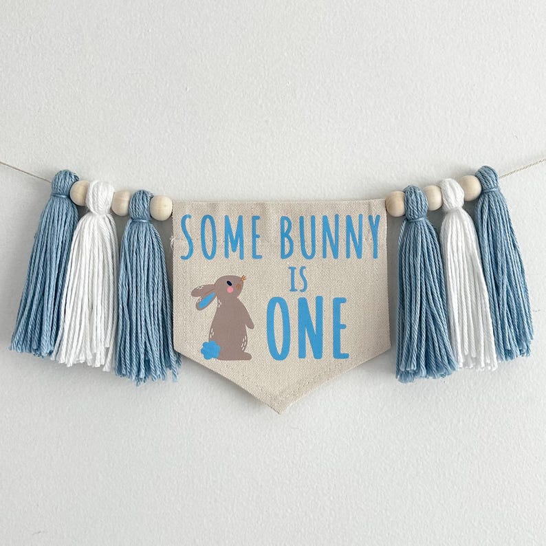 May include: A blue and white tassel garland with a banner that reads "Some Bunny is One". The banner features a brown bunny with blue accents.