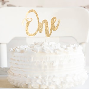 A white cake with a gold glitter One cake topper on it.