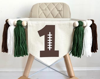 Pode incluir: Bandeira para cadeira alta com um grande número 1 castanho com costuras de futebol em tecido creme. Adornada com contas de madeira e borlas em castanho, branco e verde, ideal para uma festa de primeiro aniversário.
