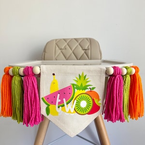 A 5 by 7 inch canvas center flag with a bunch of tropical fruit and the word TWO. There is a 5 inch hot pink, lime green and orange tarn tassel on either side, each separated by a wooden bead for a total of 6 tassels. It is hung on a high chair.
