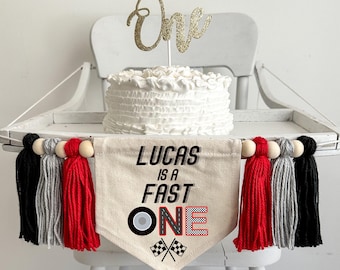 May include: A white cake with a gold glitter "One" cake topper sits on a high chair tray. A banner reading "LUCAS IS A FAST ONE" with a tire graphic and checkered flags hangs below. The banner has red, black, and gray tassels.