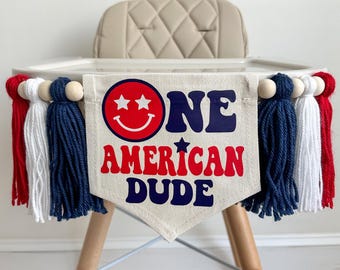 Puede incluir: Bandera para trona con el texto "ONE AMERICAN DUDE" en rojo y azul marino. La bandera tiene una carita sonriente con ojos de estrella. Está decorada con borlas rojas, blancas y azules y cuentas de madera.