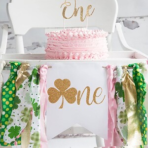 A pink birthday cake sits on a white wooden chair, decorated with a shamrock and a "One" high chair banner, surrounded by fabric ties.