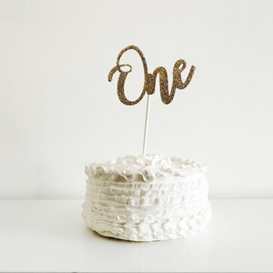 May include: A white cake with a gold glitter "One" cake topper. The cake is decorated with white frosting and has a ruffled design.