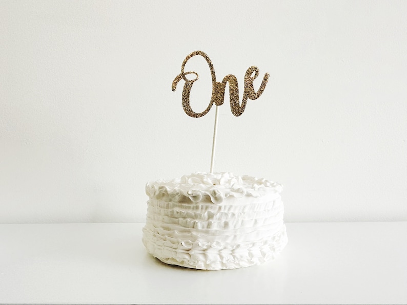 May include: A white cake with a gold glitter "One" cake topper. The cake is decorated with white frosting and has a ruffled design.