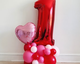 May include: A large red number one balloon with a pink heart balloon and a cluster of smaller pink and red balloons.
