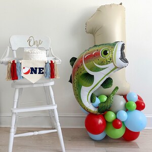 a white high chair with a cake on it, and a large balloon in the shape of a fish, along with a balloon bouquet of various colored balloons, creating a festive atmosphere.