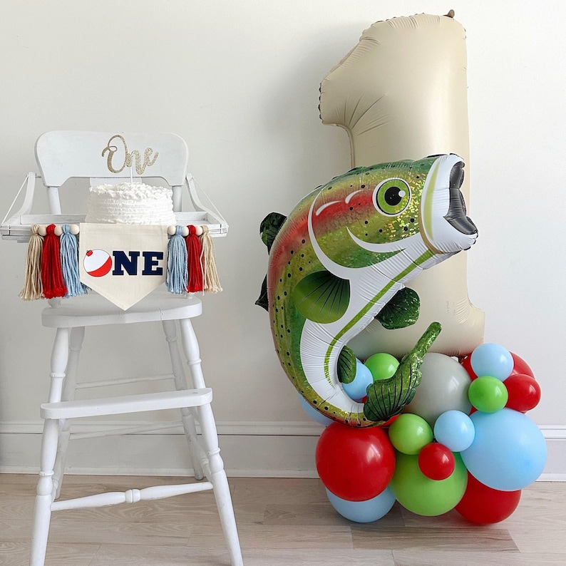 a white high chair with a cake on it, and a large balloon in the shape of a fish, along with a balloon bouquet of various colored balloons, creating a festive atmosphere.
