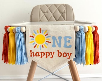 May include: A banner for a high chair with the text "ONE happy boy" and a sun graphic. The banner is decorated with red, yellow, and blue tassels and wooden beads. The high chair has a beige seat and wooden legs.