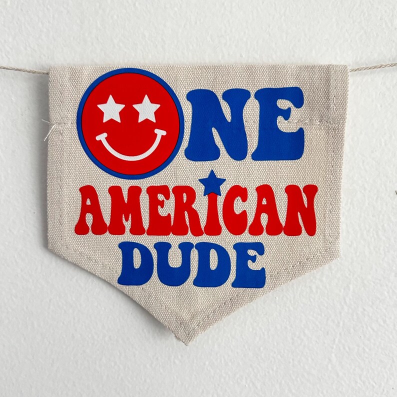 May include: A beige fabric banner with the text "ONE AMERICAN DUDE" in blue and red letters. A red smiley face with two white stars for eyes is in the top left corner.