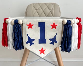 May include: A cream-colored high chair banner with red, white, and blue accents. The banner features a large blue number 1, red stars, and blue jet plane silhouettes. Tassels in red, white, and blue hang from the banner.