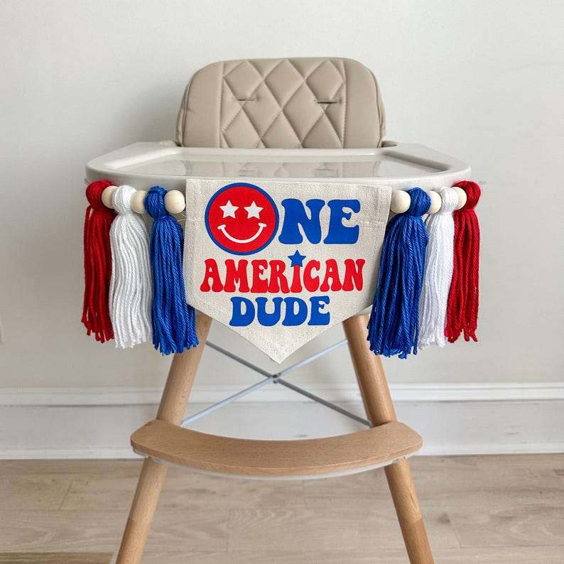 May include: A high chair with a red, white and blue banner that says "One American Dude" with a smiley face. The banner is decorated with red, white and blue tassels.