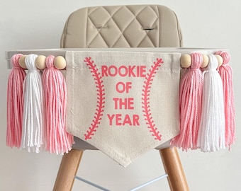 May include: A white high chair with a pink and white tassel garland and a banner that says "Rookie of the Year" with a baseball graphic.