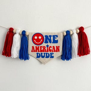 May include: A white fabric banner with red, white and blue tassels. The banner says "One American Dude" with a smiley face and stars.