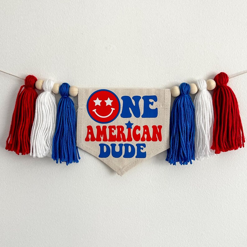 May include: A white fabric banner with red, white and blue tassels. The banner says "One American Dude" with a smiley face and stars.
