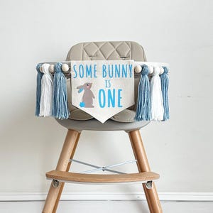 May include: A high chair with a blue and white tassel garland and a banner that says "Some Bunny Is One" with a cartoon bunny on it.
