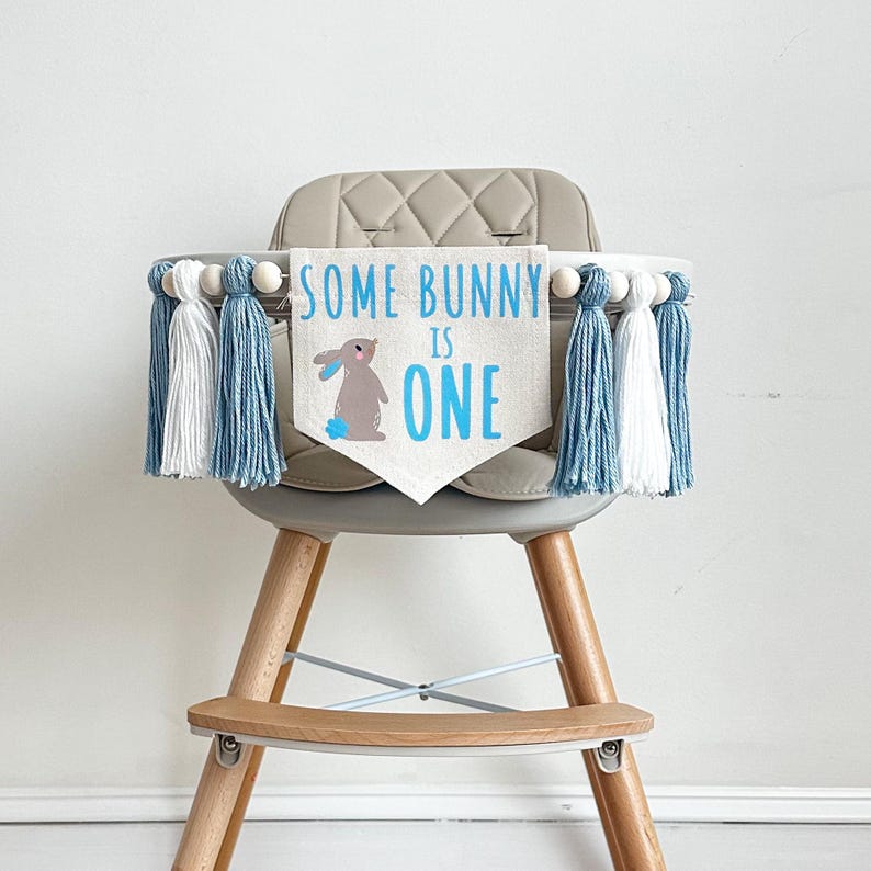 May include: A high chair with a blue and white tassel garland and a banner that says "Some Bunny Is One" with a cartoon bunny on it.