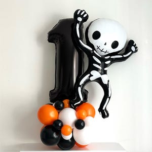 Pode incluir: Um arranjo de balões com tema de Halloween com um balão preto com o número um, um balão em forma de esqueleto e um grupo de balões laranja, pretos e brancos. O balão de esqueleto tem uma cabeça branca e um corpo preto.