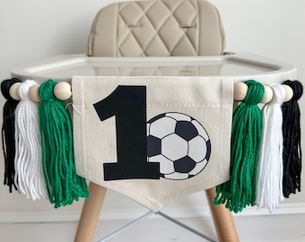 May include: A white high chair with a green, white, and black tassel garland. The garland has wooden beads and a banner that says "1" with a soccer ball.