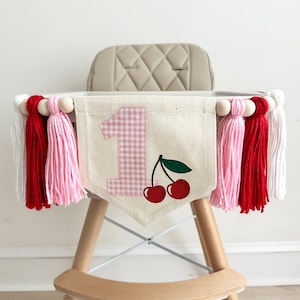 Peut inclure: Une bannière de chaise haute avec une base en toile, ornée de pompons roses et rouges, et de perles en bois. La bannière présente un numéro 1 à carreaux vichy et un motif de cerise. La chaise haute est de couleur claire avec des pieds en bois.