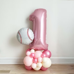 Puede incluir: Decoración de globos para una fiesta de cumpleaños. Un globo rosa con forma de número 1 y un globo de béisbol blanco con costuras rojas. Un grupo de globos rosas y blancos forma la base.