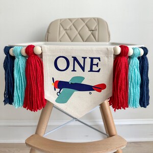 Könnte beinhalten: Ein Hochstuhlbanner mit dem Wort "ONE" in blauen Buchstaben, einem blauen und roten Flugzeug und roten, blauen und türkisfarbenen Quasten.