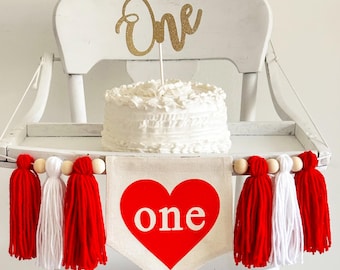 May include: White high chair with a white cake topped with a gold glitter "One" cake topper. A red heart banner with the word "one" in white letters hangs from the chair with a white and red tassel garland.