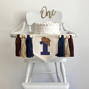 May include: A white high chair with a white cake on top. The cake is decorated with a gold glitter "one" topper. A banner with a blue number one and a brown cowboy hat hangs in front of the cake. The banner is decorated with brown, beige, and blue tassels.