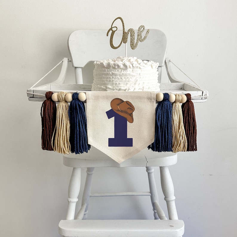 May include: A white high chair with a white cake on top. The cake is decorated with a gold glitter "one" topper. A banner with a blue number one and a brown cowboy hat hangs in front of the cake. The banner is decorated with brown, beige, and blue tassels.