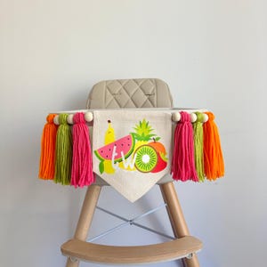 May include: A high chair banner with a fruit theme. The banner is made of burlap and has a watermelon, pineapple, kiwi, banana, and apple on it. The banner says "TWO" in white letters. The banner is decorated with colorful tassels.