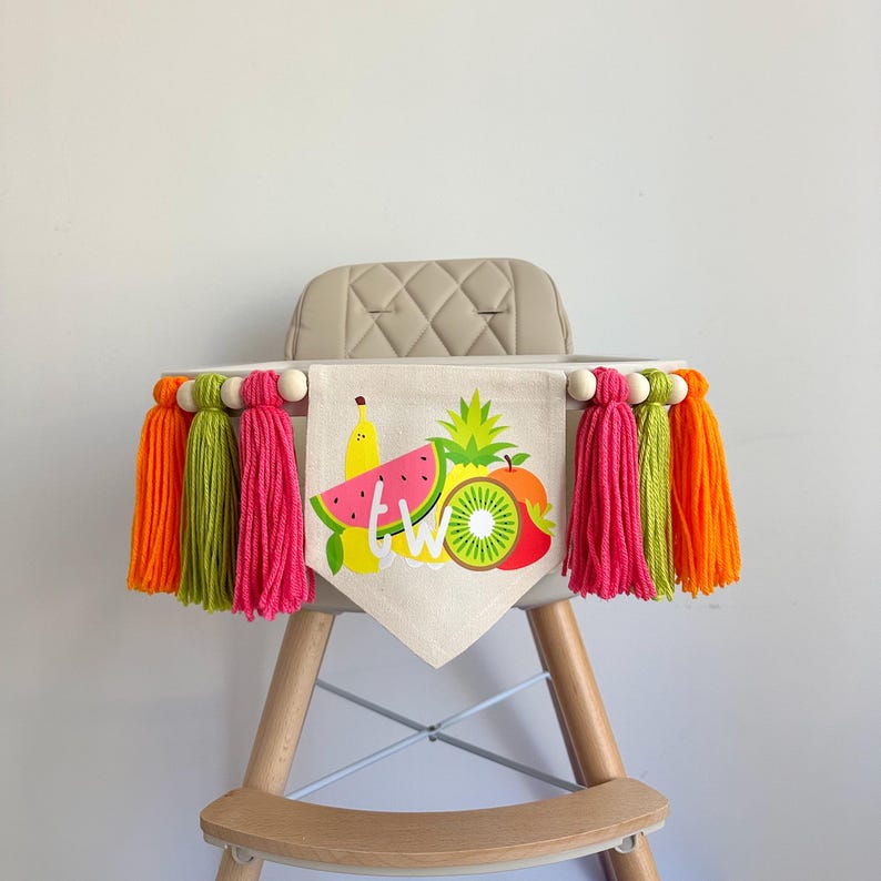 May include: A high chair banner with a fruit theme. The banner is made of burlap and has a watermelon, pineapple, kiwi, banana, and apple on it. The banner says "TWO" in white letters. The banner is decorated with colorful tassels.